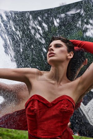 Una joven impresionante con un vestido rojo vibrante se levanta con gracia bajo la lluvia, abrazando la belleza natural del momento.