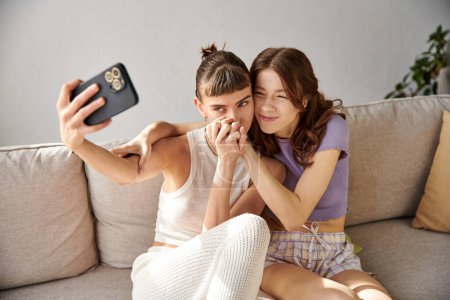 Two women in comfy attire sit on a couch, taking a selfie.