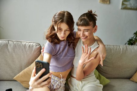 Téléchargez les photos : Couple de lesbiennes en tenue confortable assis sur le canapé, absorbé dans le téléphone portable. - en image libre de droit