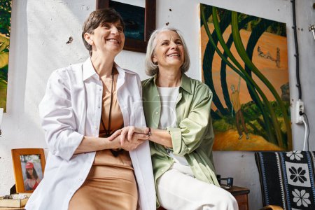 Foto de A mature lesbian couple peacefully sitting next to each other in an art studio. - Imagen libre de derechos
