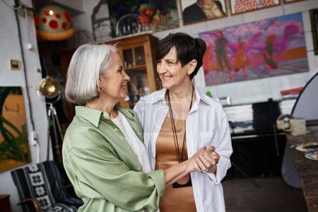 Foto de Two mature women, a lesbian couple, stand together in an art studio. - Imagen libre de derechos