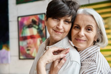 Photo for Two women hugging in front of paintings in an art studio. - Royalty Free Image