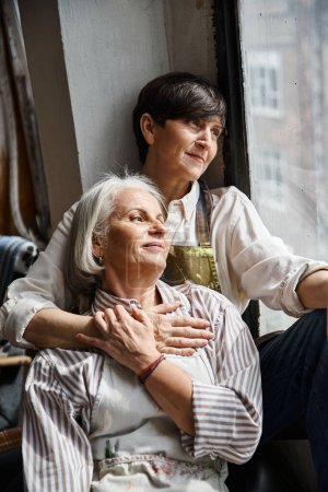 Two women sit closely on a window sill in an artists studio.