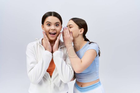 Photo for Two pretty, brunette teenage girls in sportive attire standing side by side, hands on faces, in a studio on a grey background. - Royalty Free Image