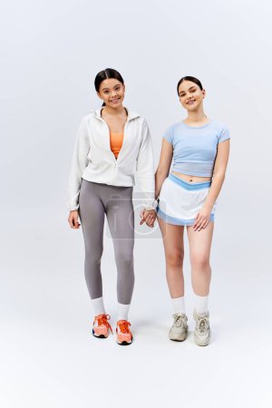 Photo for Two pretty, brunette teenage girls stand side by side, holding hands in a display of friendship on a grey studio background. - Royalty Free Image