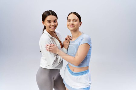 Photo for Two pretty, sportive teenage girls, one brunette, posing confidently together in a studio against a grey background. - Royalty Free Image
