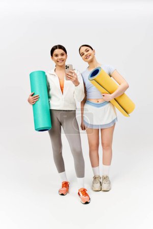 Two pretty teenage girls, one brunette, standing together in sportive attire holding yoga mats on grey studio background.
