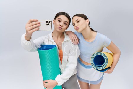 Foto de Dos guapas amigas adolescentes, luciendo cabello moreno y ropa deportiva, capturan una selfie con una esterilla de yoga en un ambiente de estudio. - Imagen libre de derechos