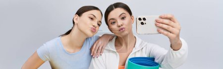 Deux jolies amies brunes en tenue sportive prenant un selfie avec un téléphone portable sur fond de studio gris.