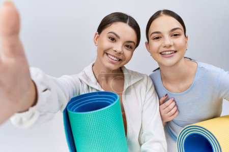 Zwei hübsche und brünette Teenager-Mädchen, Freundinnen, in sportlicher Kleidung stehen nebeneinander mit Yogamatten in einem Studio auf grauem Hintergrund.