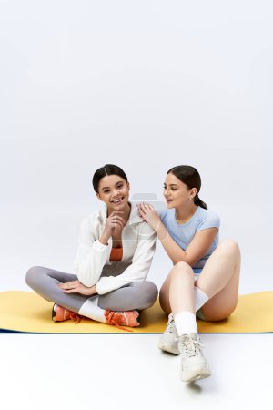 Photo for Two pretty, brunette teenage girls in sportive attire sitting on a mat, holding hands in unity and friendship. - Royalty Free Image