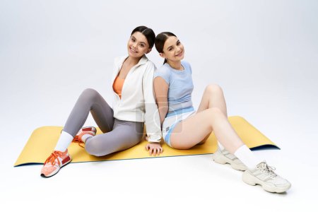 Téléchargez les photos : Deux jolies adolescentes brunes en tenue sportive s'assoient ensemble sur un tapis dans un studio, mettant en valeur l'amitié et la détente. - en image libre de droit