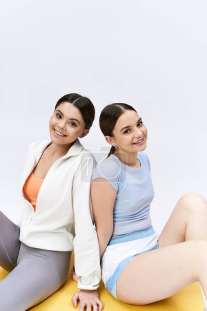 Two pretty teenage girls, brunette, in sportive attire, sit on a yellow mat against a grey background, sharing a fun moment together.