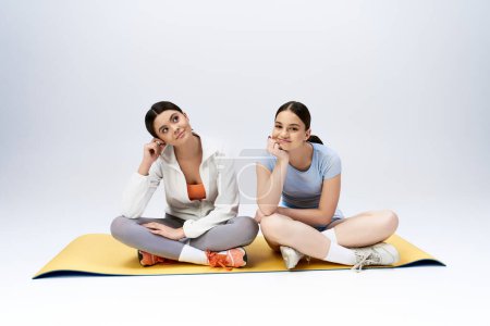 Foto de Dos guapas adolescentes morenas en traje deportivo se sientan en una esterilla de yoga, compartiendo un momento de relajación y conexión. - Imagen libre de derechos