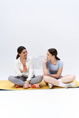 Photo for Two pretty, brunette teenage girls in sportive attire sit on a yoga mat, talking and connecting in a serene studio setting. - Royalty Free Image