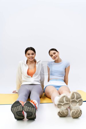 Zwei hübsche und brünette Teenager-Mädchen in sportlicher Kleidung sitzen zusammen auf einer Matte mit den Füßen in einem Studio-Setting.