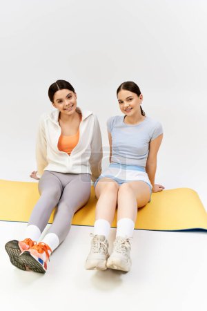 Photo for Two pretty and brunette teenage girls in sportive attire sitting on a mat with legs crossed in a studio. - Royalty Free Image