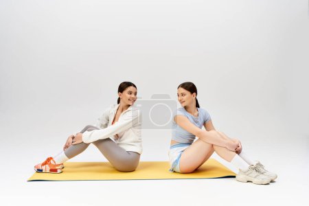 Foto de Dos guapas adolescentes morenas con atuendo deportivo comparten un momento sereno mientras se sientan en una esterilla de yoga en un estudio. - Imagen libre de derechos