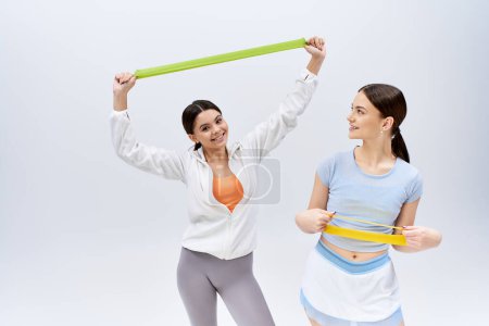 Two pretty teenage brunette girls in sportive attire stand as female friends in a studio against a grey background.