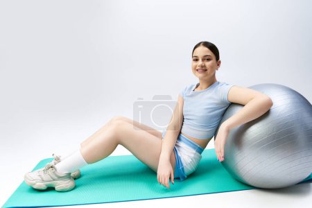 A pretty, brunette girl in sportive attire sits gracefully on top of a gym ball in a studio setting