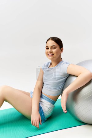 A pretty, brunette teenage girl in sporty attire gracefully balances on a yoga ball in a studio