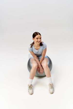 A young girl, wearing sportive attire, elegantly sits atop a fitness ball in a studio setting.