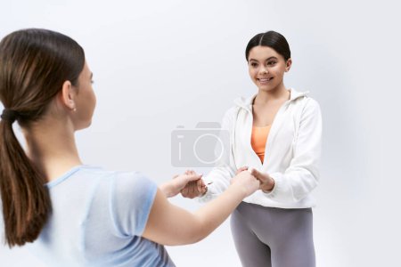 Two pretty brunette teenage girls in sportive attire hold hands, embodying friendship and trust on a grey studio background.