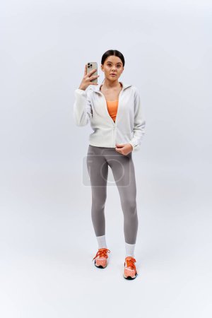 Téléchargez les photos : Une fille brune en tenue de sport prend joyeusement un selfie avec son téléphone portable dans un cadre studio. - en image libre de droit