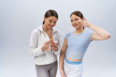 Téléchargez les photos : Deux jolies adolescentes brunes en tenue de sport se tiennent côte à côte avec une pose confiante sur un fond de studio gris. - en image libre de droit