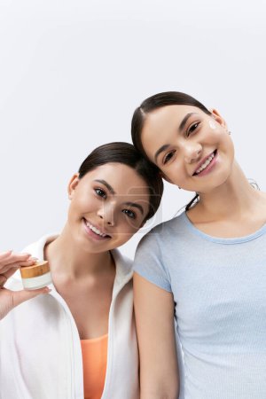Zwei hübsche Mädchen im Teenageralter, beide mit brünetten Haaren, stehen zusammen in einem Studio vor grauem Hintergrund und lächeln fröhlich..