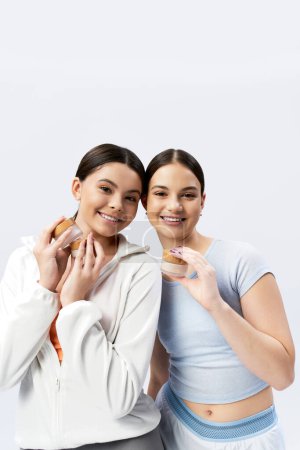 Two pretty, brunette teenage girls holding cream on a grey studio background, showing friendship.