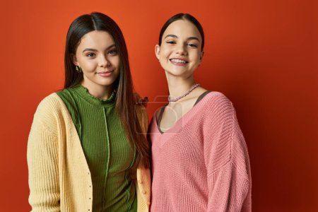 Foto de Dos chicas adolescentes guapas y morenas en atuendo casual paradas una al lado de la otra frente a una vibrante pared roja. - Imagen libre de derechos