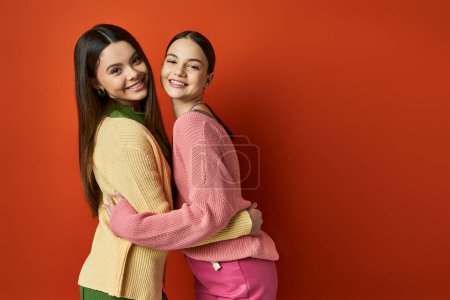 Téléchargez les photos : Deux jeunes femmes élégantes avec les cheveux bruns et tenues décontractées debout côte à côte devant un mur rouge vibrant. - en image libre de droit