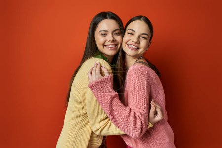 Foto de Dos guapas adolescentes morenas con atuendo casual abrazándose frente a una vibrante pared roja. - Imagen libre de derechos