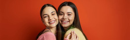 Photo for Two young women, pretty and brunette, hug each other affectionately in front of a vibrant red wall. - Royalty Free Image