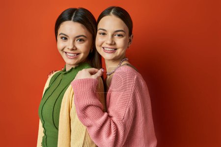 Foto de Dos chicas adolescentes guapas, vestidas casualmente, morenas se paran lado a lado frente a una pared roja vívida, simbolizando la amistad. - Imagen libre de derechos