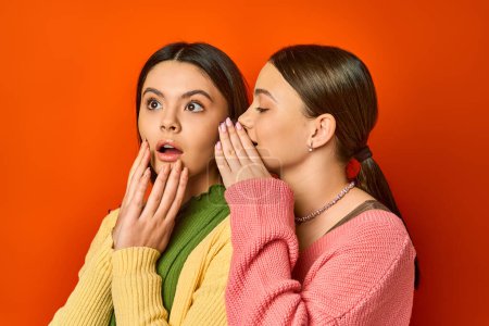 Two pretty, brunette teenage girls in casual attire stand next to each other with mouths open in surprise on an orange studio background.