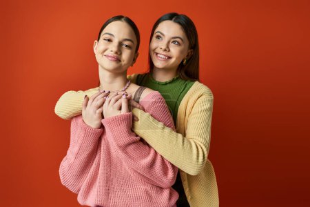 Two pretty, brunette teenage girls hug each other warmly in front of a bold red background, showcasing their deep friendship.