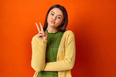 A pretty brunette teenage girl makes a peace sign with her fingers in a studio on an orange background.