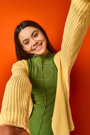 Photo for A brunette teenage girl wearing a green dress and a yellow cardigan poses elegantly on an orange background in a studio. - Royalty Free Image