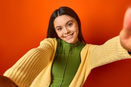 A pretty, brunette teenage girl smiles, wearing a green shirt and a yellow cardigan, posing on an orange background.