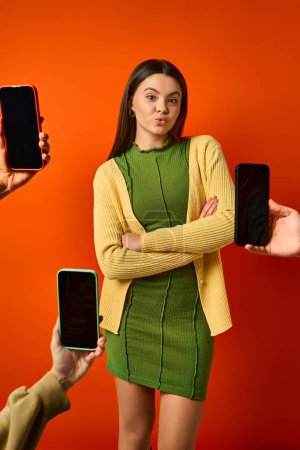 Ein brünettes Teenager-Mädchen in einem grünen Kleid in der Nähe von Mobiltelefonen, umgeben von ihr auf orangefarbenem Hintergrund in einem Studio.