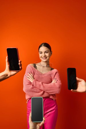 Ein hübsches, brünettes Teenager-Mädchen steht vor einer leuchtend roten Wand in der Nähe von Mobiltelefonen und präsentiert moderne Verbindung und Technologie..