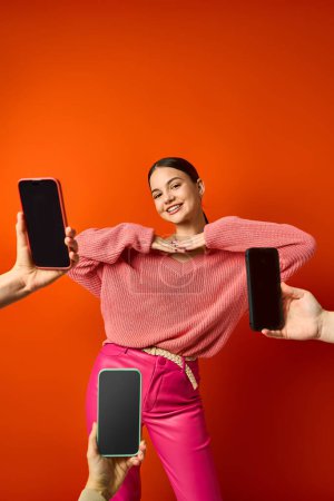 A brunette teenage girl in pink near cell phones against an orange background in a studio.