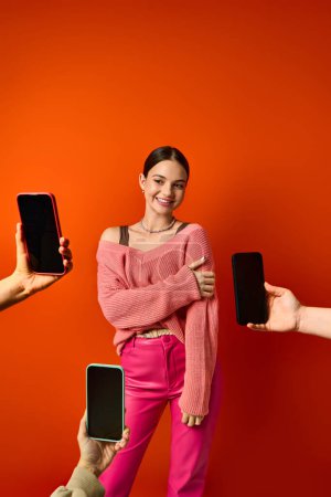 A brunette woman stands before a red wall, near two cell phones in hands of people
