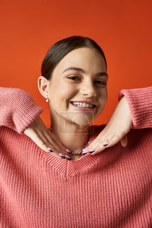 A brunette teenage girl in a pink sweater is smiling brightly against an orange background in a studio setting.