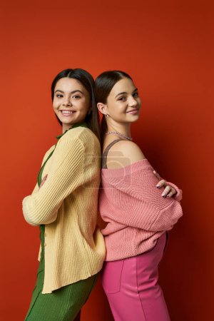Foto de Dos guapas adolescentes morenas con atuendo casual paradas una al lado de la otra frente a una vibrante pared roja. - Imagen libre de derechos