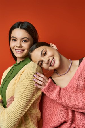 Two pretty, brunette teenage girls wearing casual attire hugging each other in front of a vivid red wall in a studio.