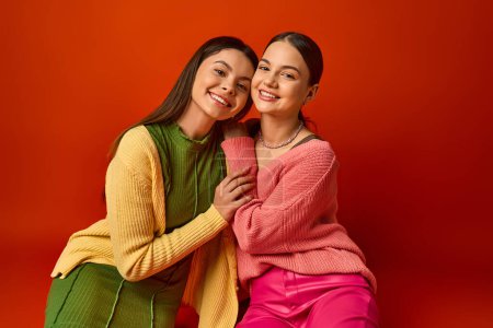 Photo for Two pretty brunette teenage girls in casual attire sitting next to each other on a vibrant red background in a studio setting. - Royalty Free Image