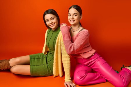 Téléchargez les photos : Deux jolies adolescentes brunes s'assoient par terre en tenue décontractée, prenant une pose pour une photo sur fond orange. - en image libre de droit